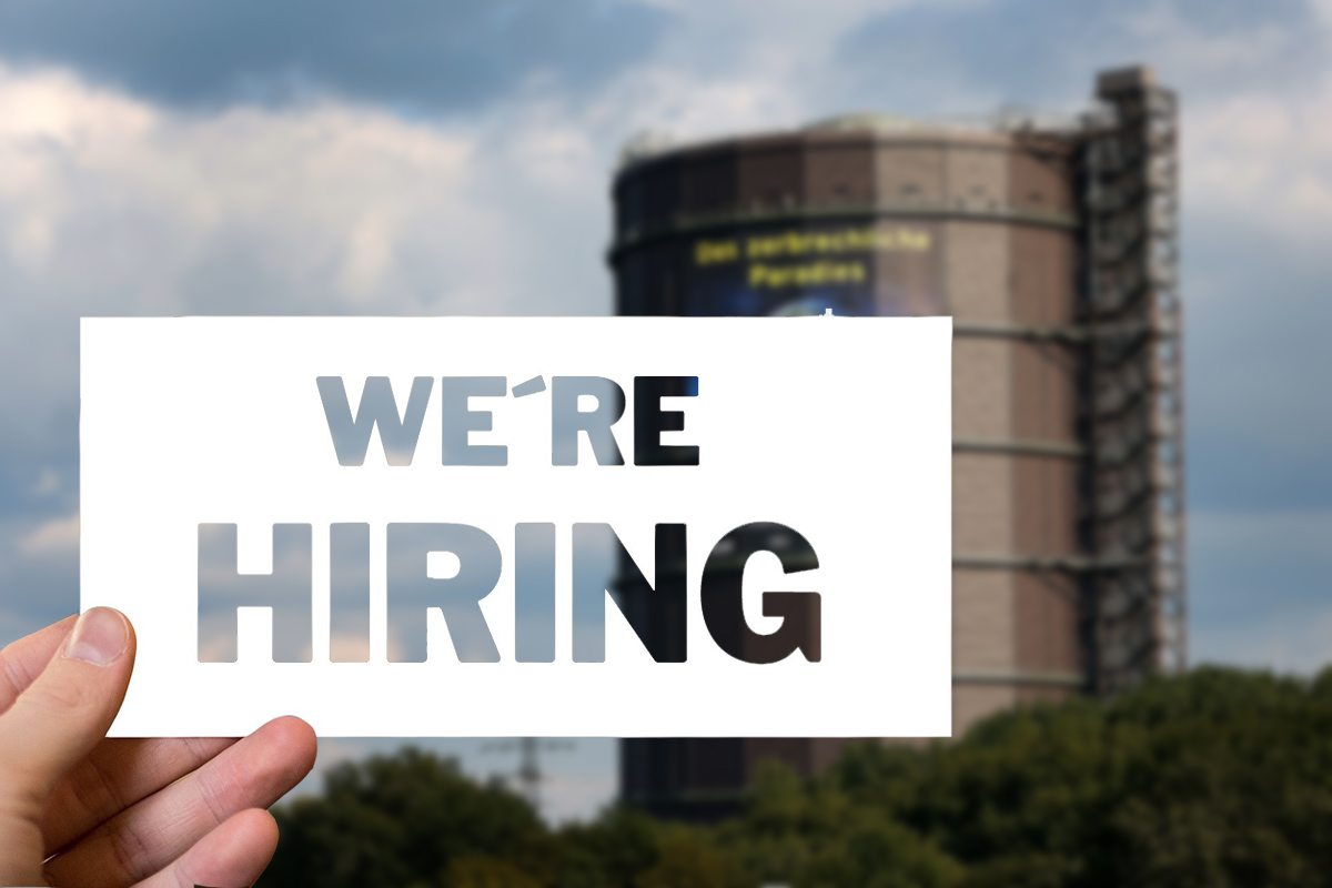Ein AHnd hält ein Schild mit dem Text: We are hiring. Im Hintergrund ist der unscharfe Gasometer zu sehen.