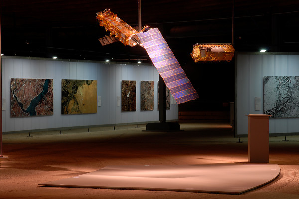 Ein Satellit hängt vor verschiedenen großformatigen Ausstellungsbildern
